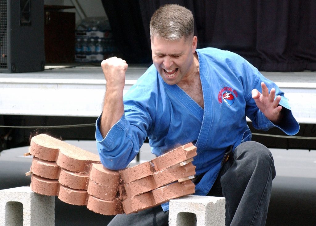 Karateka in a blue uniform using a downward elbow strike to break a stack of three clay tiles, each about eight centimetres thick